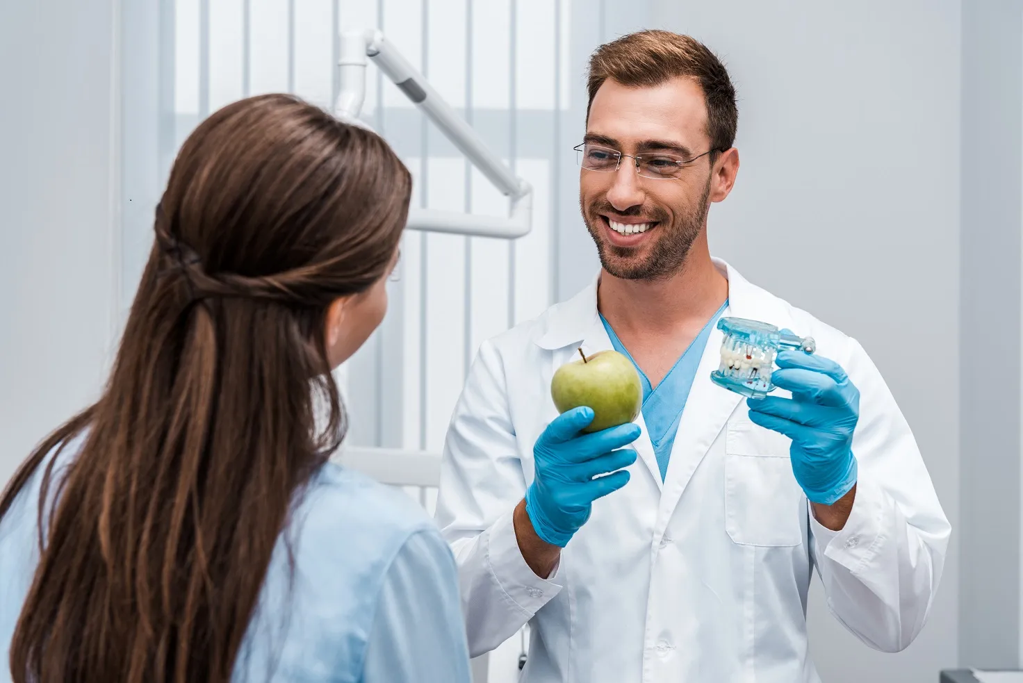 selective focus of happy dentist in glasses holding tooth model near patient